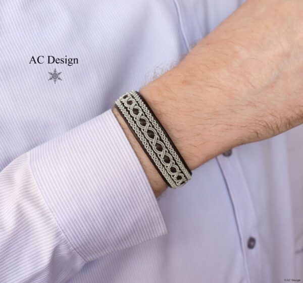 A sami bracelet is shown on a mans wrist. The man wears a light skirt with stripes. The company name AC Design is written in black with a star below.